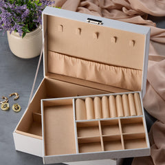 A white jewelry box with multiple compartments, placed on a table next to a vase of purple flowers.