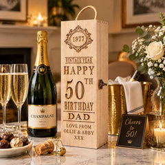a wooden box with a personalized message, a bottle of champagne, and other celebratory items such as a vase of flowers, a candle, and a bowl of chocolates.