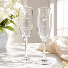 two clear glass goblets with personalized messages on them, placed on a table with a vase of white flowers in the background.