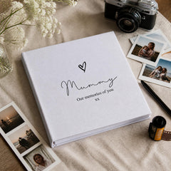 A white photo album with the text "Mummy Our memories of you" printed on it, surrounded by photographs, a camera, and other personal items on a table.