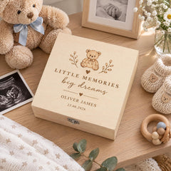 A wooden box with a personalized message, a teddy bear, and other baby-related items on a wooden table.