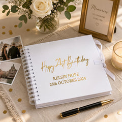 a white notebook with gold text and a pen, along with a framed picture, a vase of flowers, and some confetti on a table.