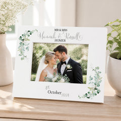 A white picture frame with a photo of a bride and groom inside, surrounded by green leaves and flowers. The frame has a decorative border and the names "Mr & Mrs" and "Hannah & Russell" written on it.
