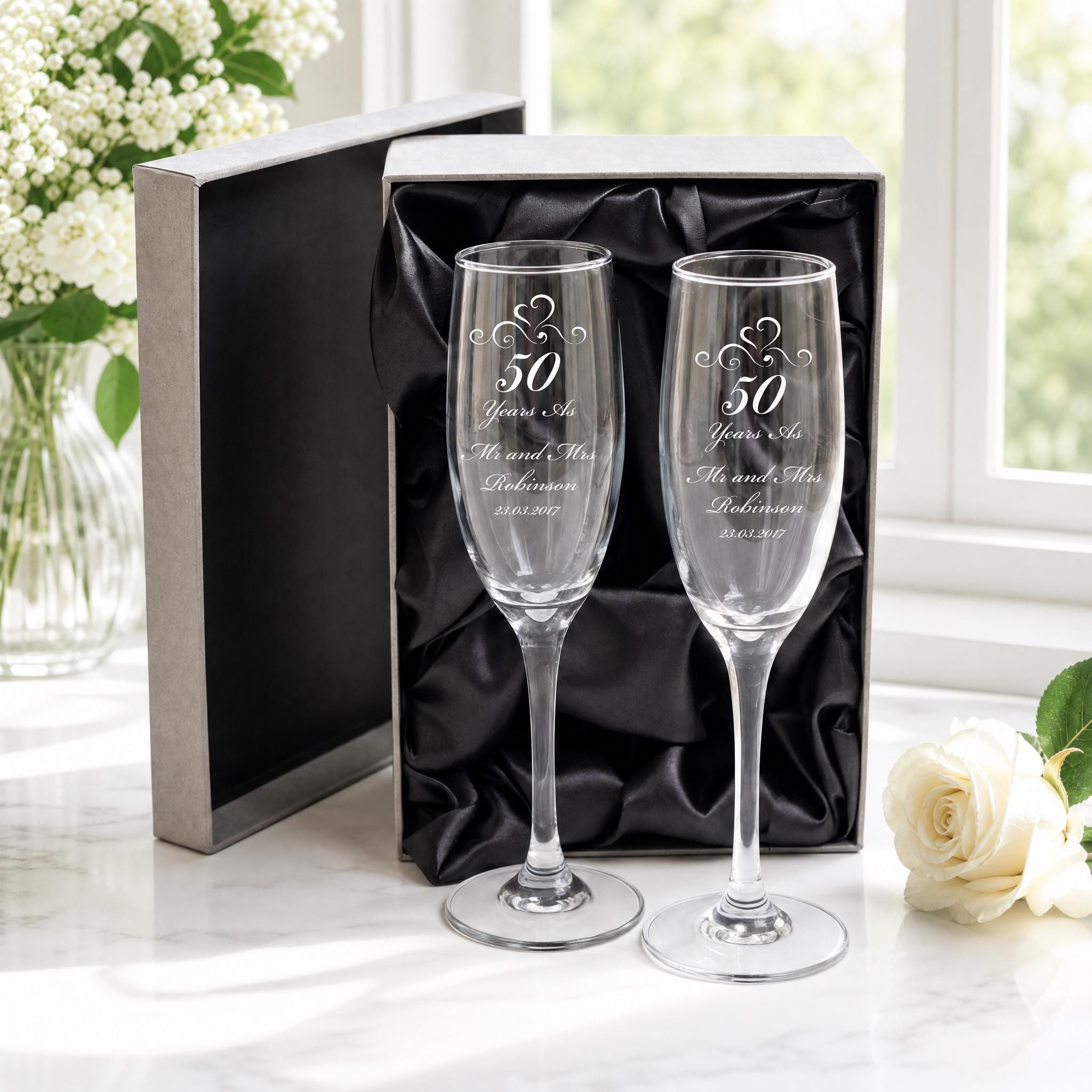 a pair of elegant champagne glasses, each inscribed with "50 Years As Mr & Mrs Robinson" and "26/03/2017", placed on a white marble surface. The glasses are accompanied by a black box and a vase of white flowers, creating a sophisticated and romantic atmosphere.