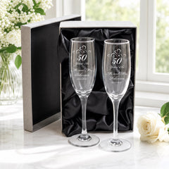 a pair of elegant champagne glasses, each inscribed with "50 Years As Mr & Mrs Robinson" and "26/03/2017", placed on a white marble surface. The glasses are accompanied by a black box and a vase of white flowers, creating a sophisticated and romantic atmosphere.