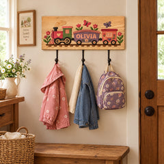 A wooden coat rack with three hooks, displaying a train-themed coat rack with a personalized name, a pink coat, a blue denim jacket, and a purple backpack.