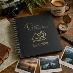 a black spiral-bound notebook with the text "Our Adventures Tom & Ashleigh" printed on it, along with a compass and a mug labeled "Adventure Awaits". The notebook is surrounded by various travel-related items, including a map, photographs, and a pen.
