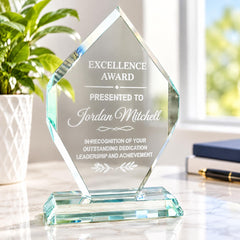 A clear glass award trophy with an inscription, placed on a marble surface next to a potted plant.