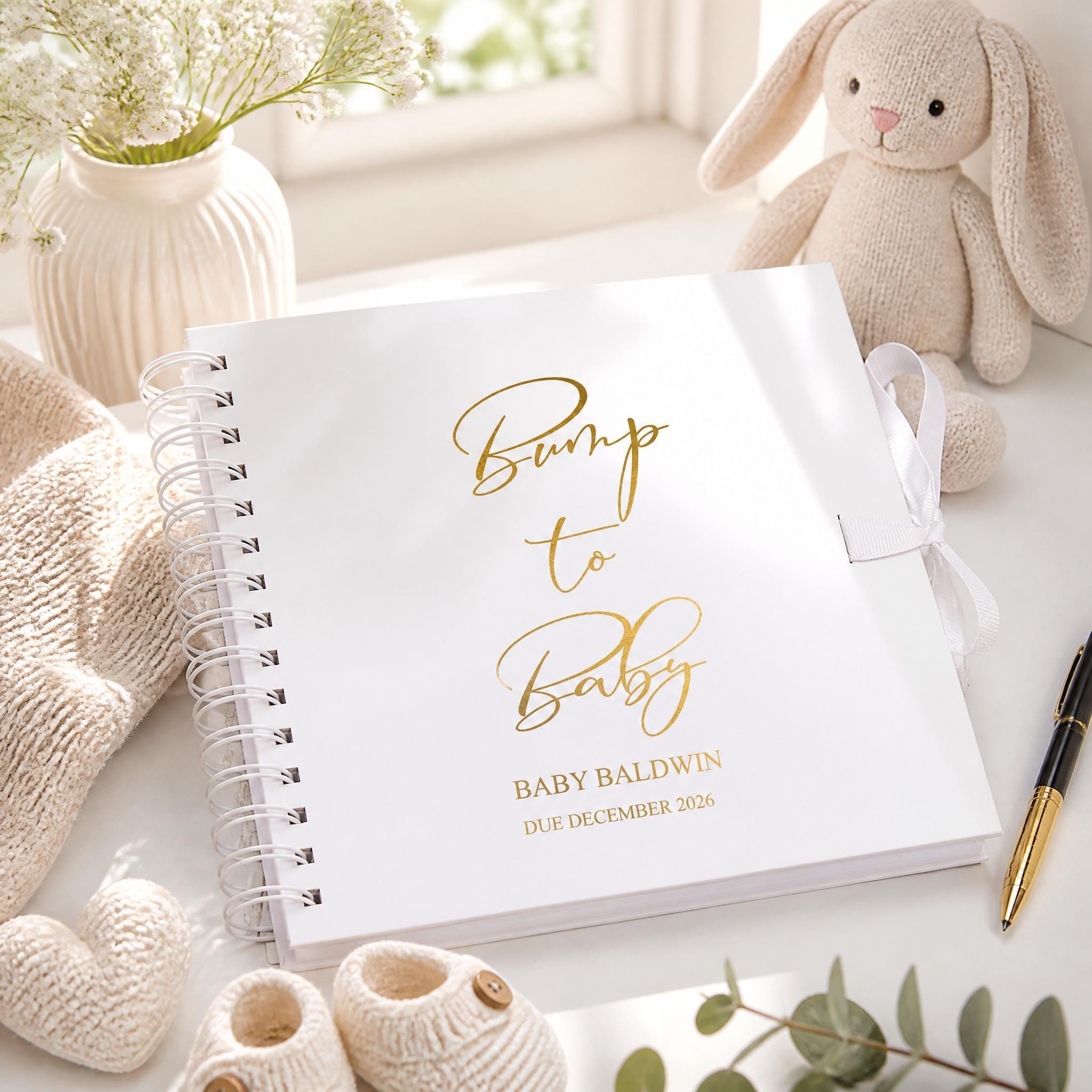 a white spiral-bound book titled "Bump to Baby" with gold lettering, placed on a white surface next to a stuffed rabbit, a vase with flowers, and a pen.