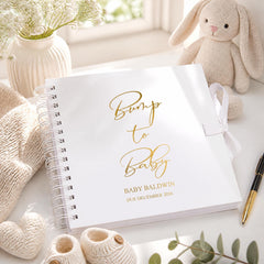 a white spiral-bound book titled "Bump to Baby" with gold lettering, placed on a white surface next to a stuffed rabbit, a vase with flowers, and a pen.