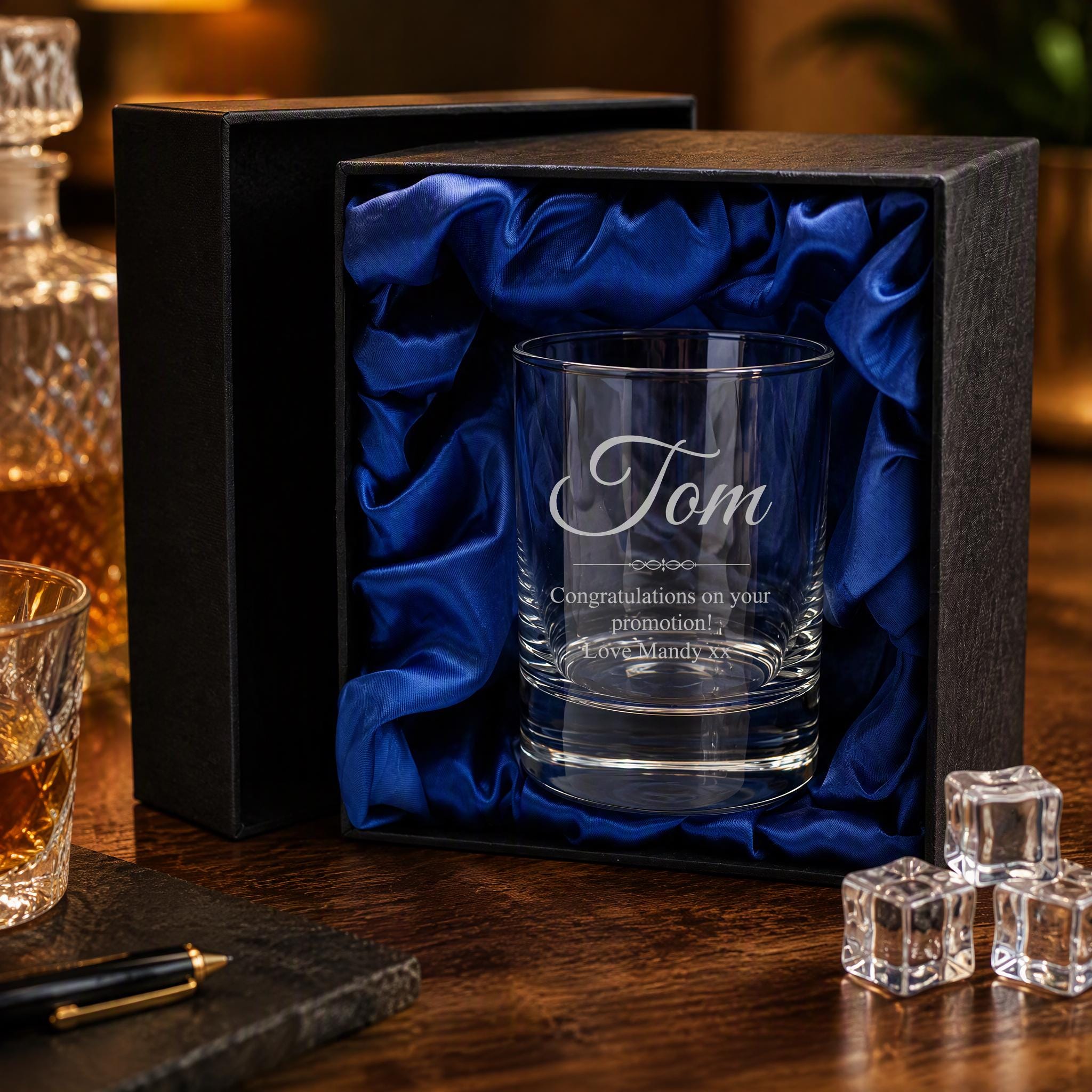 A personalized glass with the name "Tom" engraved on it, placed in a black box with a blue satin lining. The glass is accompanied by a pen and some ice cubes, suggesting a gift or a special occasion.