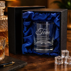 A personalized glass with the name "Tom" engraved on it, placed in a black box with a blue satin lining. The glass is accompanied by a pen and some ice cubes, suggesting a gift or a special occasion.