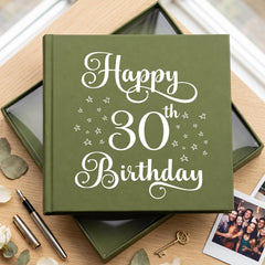 A green photo album with the text "Happy 30th Birthday" printed on it, placed on a wooden surface alongside some flowers and a key.
