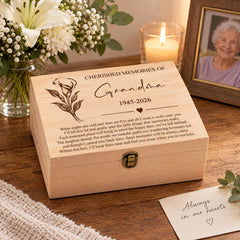 a wooden box with a heartfelt message engraved on it, placed on a wooden surface. The box is accompanied by a note that reads "Always in our hearts". In the background, there is a vase filled with white flowers, and a framed photograph of an elderly woman.