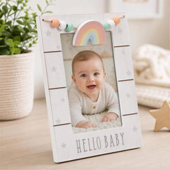 A baby smiling in a white wooden frame with a rainbow and star design, placed on a wooden surface next to a potted plant.