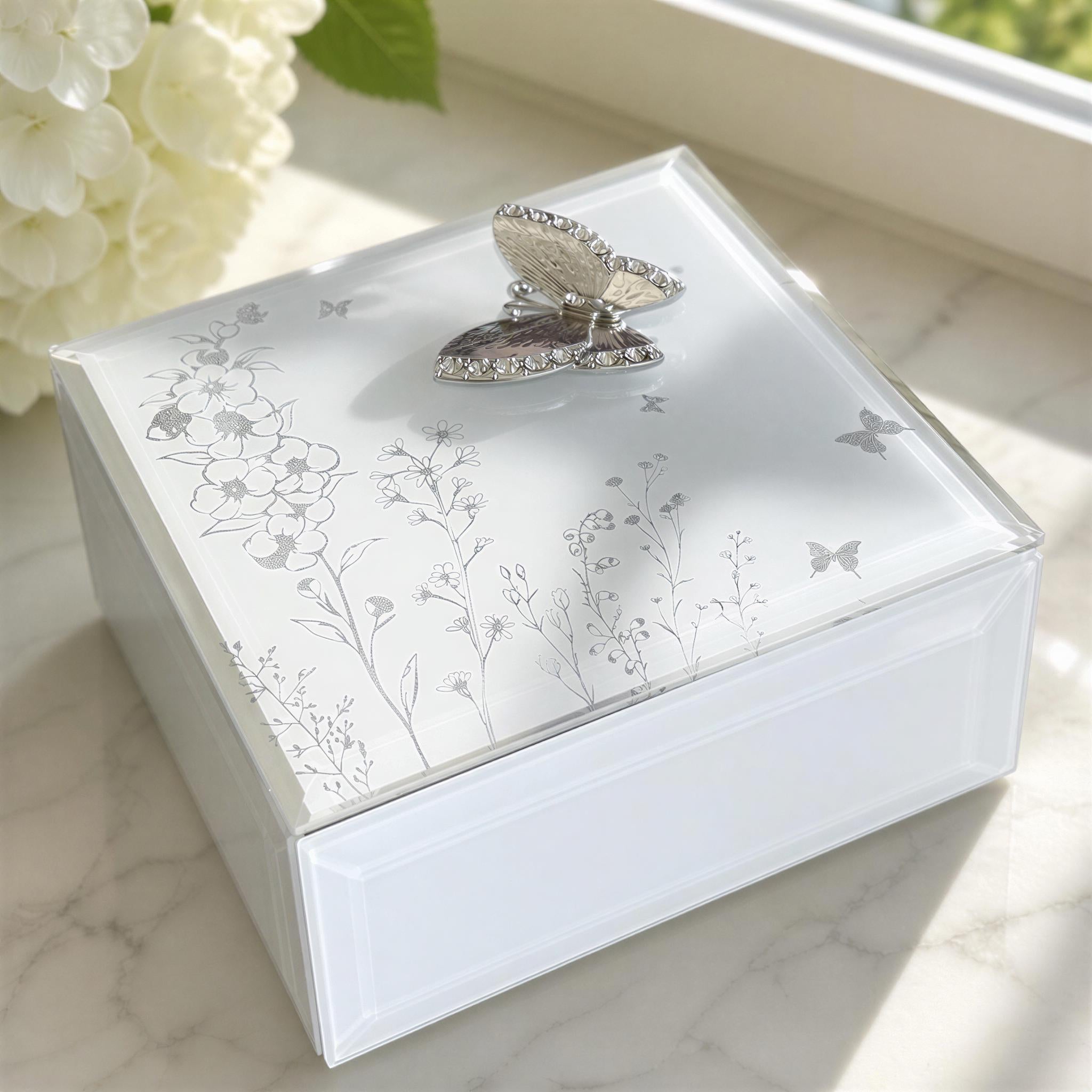 A white, square-shaped box with a silver butterfly design on the lid, placed on a marble surface near a window.