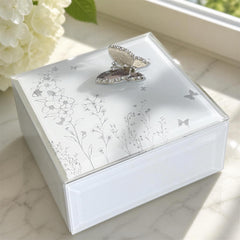 A white, square-shaped box with a silver butterfly design on the lid, placed on a marble surface near a window.