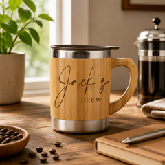 A wooden mug with the text "Jack's BREW" engraved on it, placed on a wooden surface next to a coffee pot, a notebook, and some coffee beans.