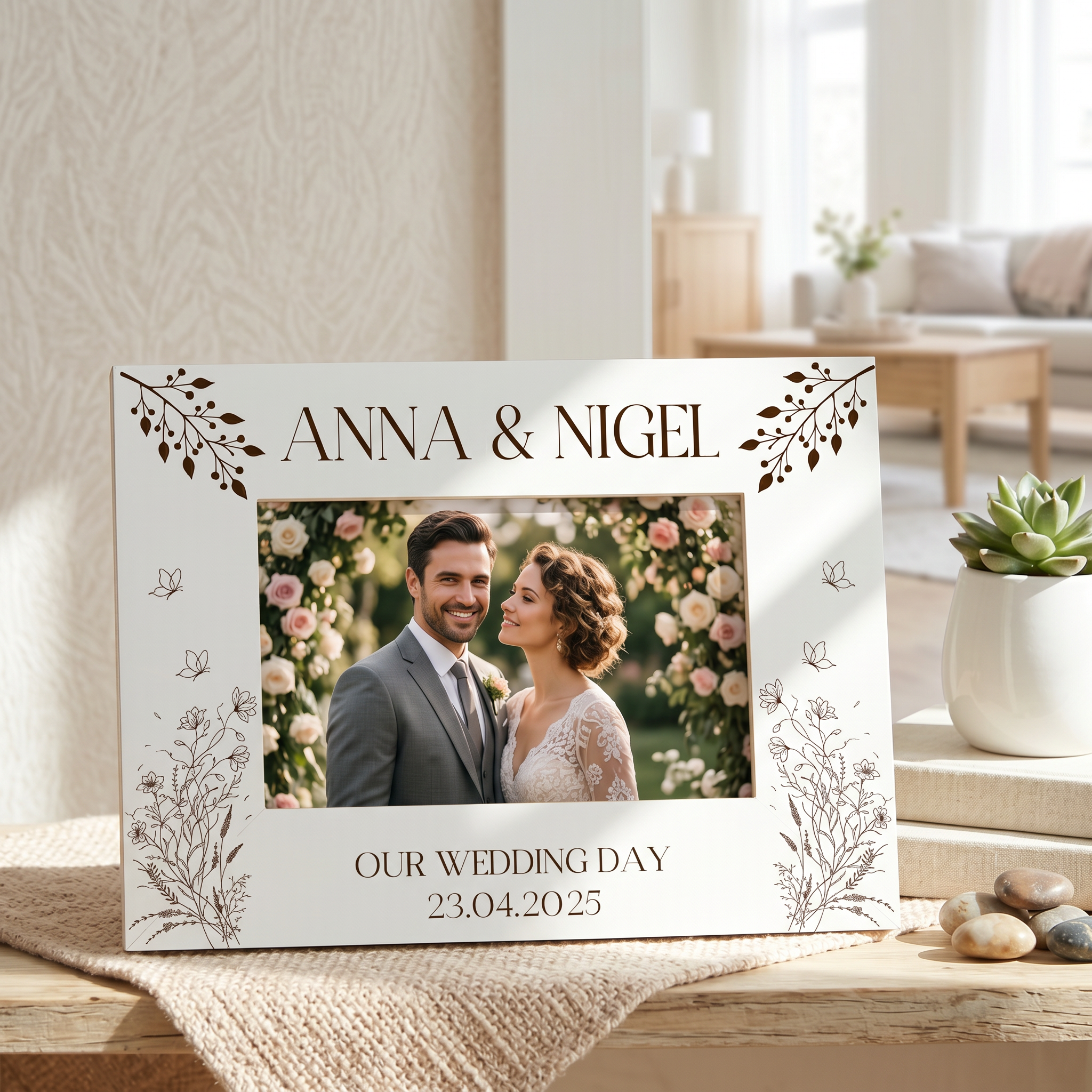 A white picture frame with a photo of a couple, surrounded by decorative floral elements, sits on a wooden surface. The frame has the text "ANNA & NIGEL" and "OUR WEDDING DAY 23.04.2025" printed on it.