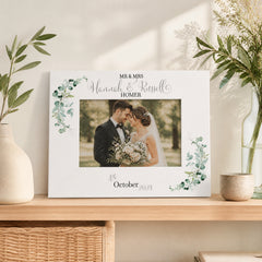 A white picture frame with a photo of a couple, surrounded by green leaves, sits on a shelf. The frame has text that reads "Mr & Mrs Hannah & Russell" and "October 2034".
