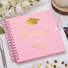 A pink graduation yearbook with gold text and a graduation cap sits on a marble surface, surrounded by white flowers, a pen, and a wooden coaster.
