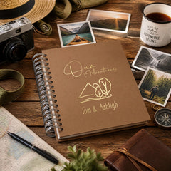 a brown notebook with the title "Our Adventures Tom & Ashleigh" on it, surrounded by various travel-related items such as a camera, a hat, a compass, and a mug.
