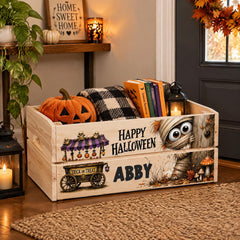 A wooden crate decorated for Halloween, featuring a carved pumpkin, a ghost, and a skeleton. The crate has a sign that says "Happy Halloween Abby" and is placed on a rug.