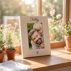 A white picture frame with a photo of a smiling man and a young boy, placed on a windowsill with potted plants in the background.