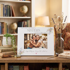 a white picture frame with a photo of two smiling women, placed on a wooden surface next to a vase of dried flowers and a stack of books.