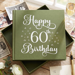 A green book titled "Happy 60th Birthday" sits on a wooden table, surrounded by family photos, a wine glass, and a note.