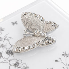 A silver butterfly brooch with rhinestones on it, resting on a white surface with a floral pattern.