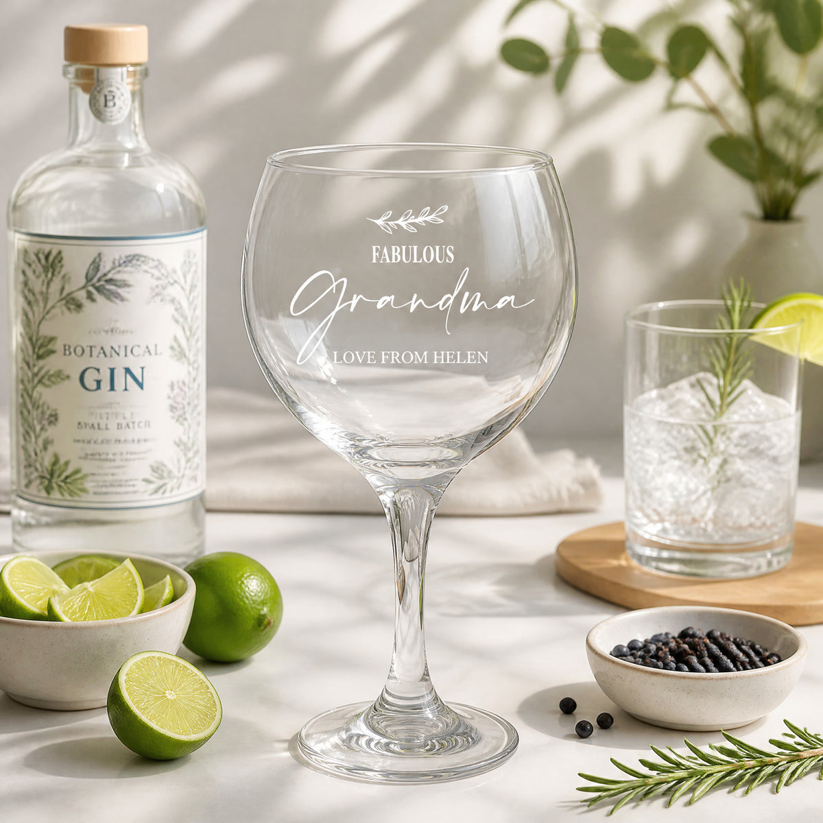 a table with a bottle of Botanical Gin, a wine glass with a personalized message, a glass of water, a bowl of black peppercorns, and some lime wedges.