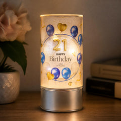 A birthday-themed lighted lamp with a design of blue and gold balloons and the text "Happy Birthday" on it, placed on a wooden surface next to a potted plant.
