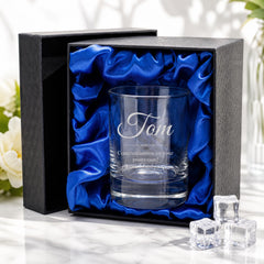 A personalized glass with the name "Tom" engraved on it, placed in a black box with a blue satin lining. The glass is surrounded by ice cubes, and there are some flowers in the background.