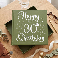 A green book titled "Happy 30th Birthday" sits on a wooden table, surrounded by various items such as a wine glass, glasses, a pen, and a ribbon.