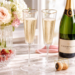 a bottle of champagne, two champagne glasses filled with champagne, a vase of white flowers, and a bowl of strawberries on a marble countertop.