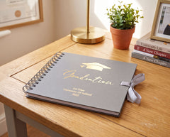 a graduation album on a wooden desk, with a potted plant and books in the background.