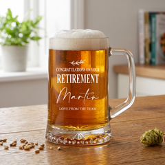 A glass mug filled with beer, placed on a wooden surface, with a potted plant in the background.