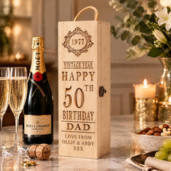 A wooden box with a personalized message for a 50th birthday celebration, featuring a champagne bottle and glasses, as well as a bowl of nuts and a vase of flowers.