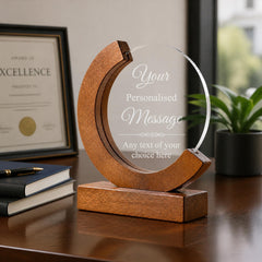 A wooden award or plaque with a clear glass front, placed on a wooden desk. The plaque has a message that reads "Your Personalised Message Any text of your choice here". In the background, there is a certificate and a potted plant.