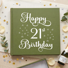 a green book with the text "Happy 21st Birthday" written on it, surrounded by confetti, a bottle of champagne, and some leaves.
