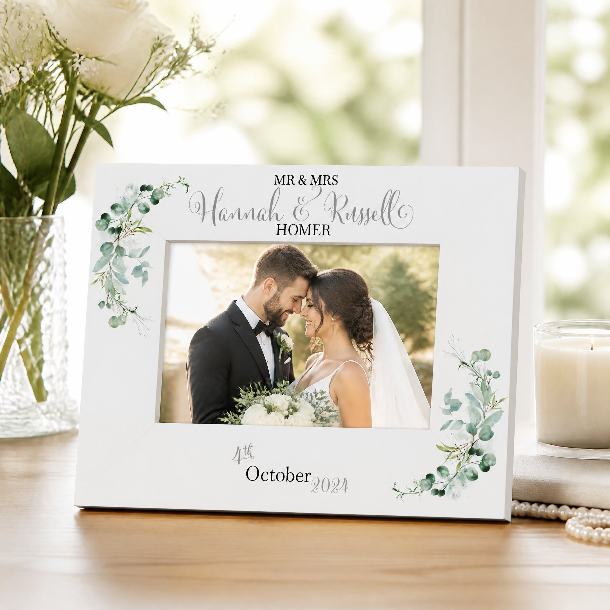 A white picture frame with a photo of a bride and groom, surrounded by floral decorations, sits on a wooden surface.
