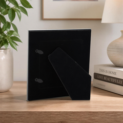 a black picture frame with a plant in a white vase on a wooden surface, with a stack of books and a lamp in the background.