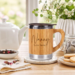 A wooden mug with the name "Nanny" and the name "Love Jamie" printed on it, sitting on a wooden table next to a white teapot, a plate of cookies, and a vase with flowers.