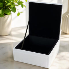 a white, square-shaped box with a black interior, placed on a marble surface. There is a plant in the background, adding a touch of greenery to the scene.