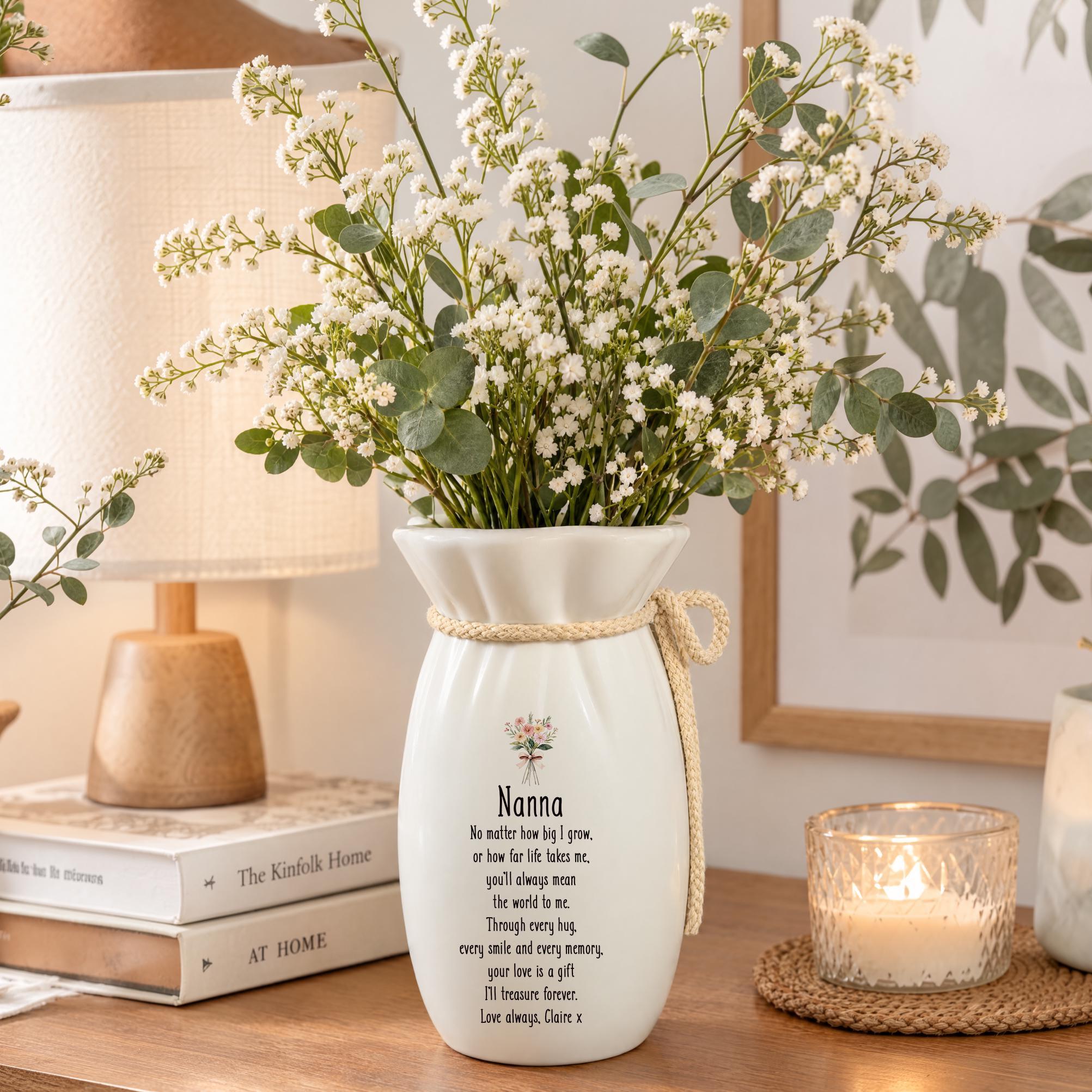 A white vase with a bouquet of white flowers sits on a wooden surface, accompanied by a stack of books and a candle.
