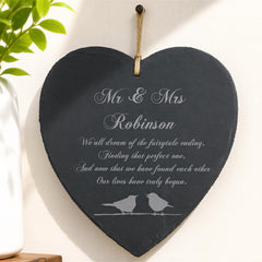 A heart-shaped sign with a couple's names and a romantic message, hanging on a wall next to a plant.