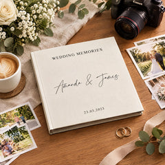 A wedding album titled "Wedding Memories" with the names "Amanda & James" printed on it, placed on a wooden surface alongside wedding photos, a camera, and a cup of coffee.