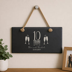 A black slate sign with a white design hangs on a wall, celebrating a 10-year anniversary with two champagne glasses and the text "10 YEARS TOGETHER" and "Ben & Rachel 23.03.2019".