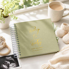 a baby shower gift set, including a green notebook with gold text, a white mug with a heart design, a baby bootie, and a black and white photograph of an infant.